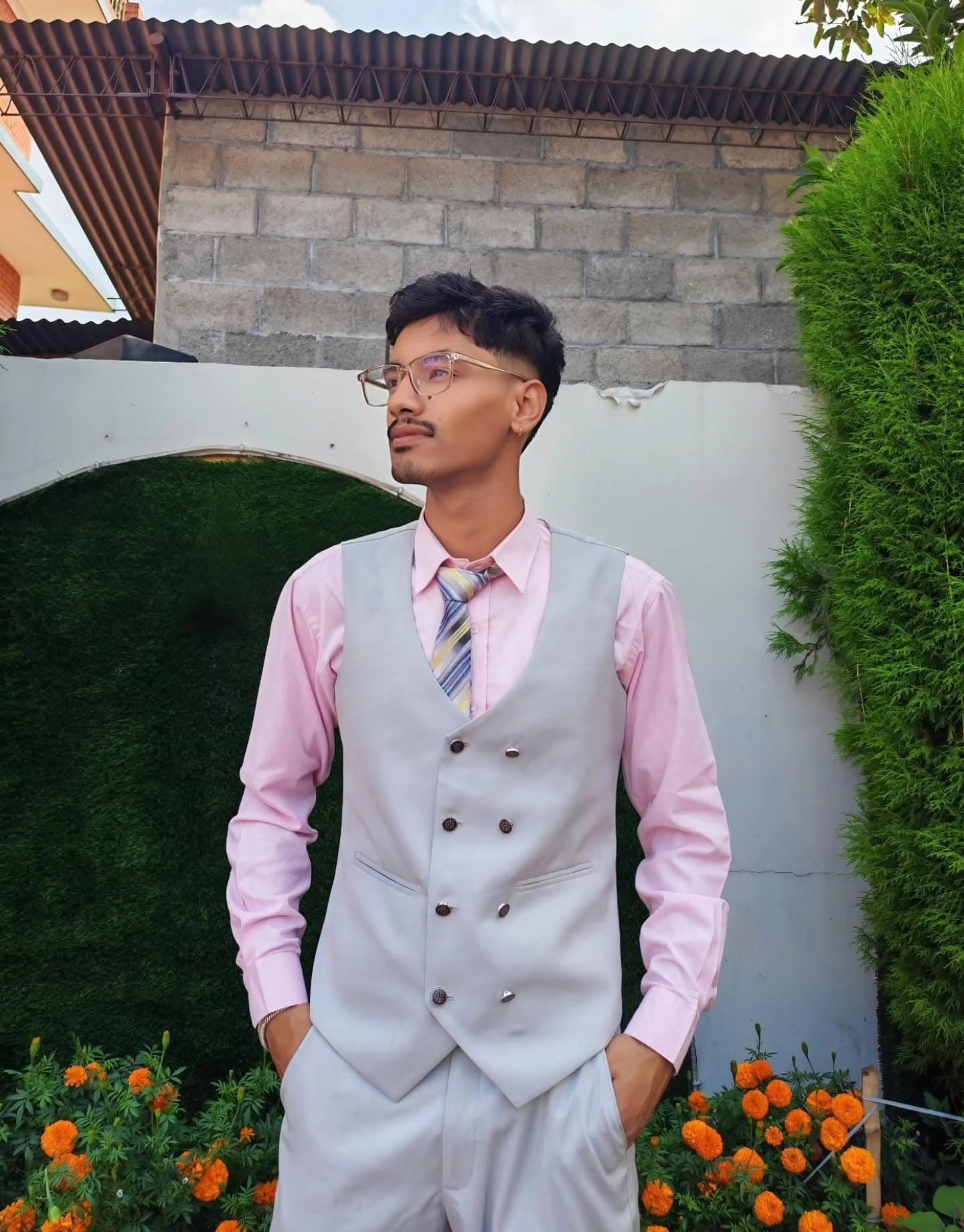 Young man in a pink shirt and grey waistcoat posing outdoors near a brick wall.