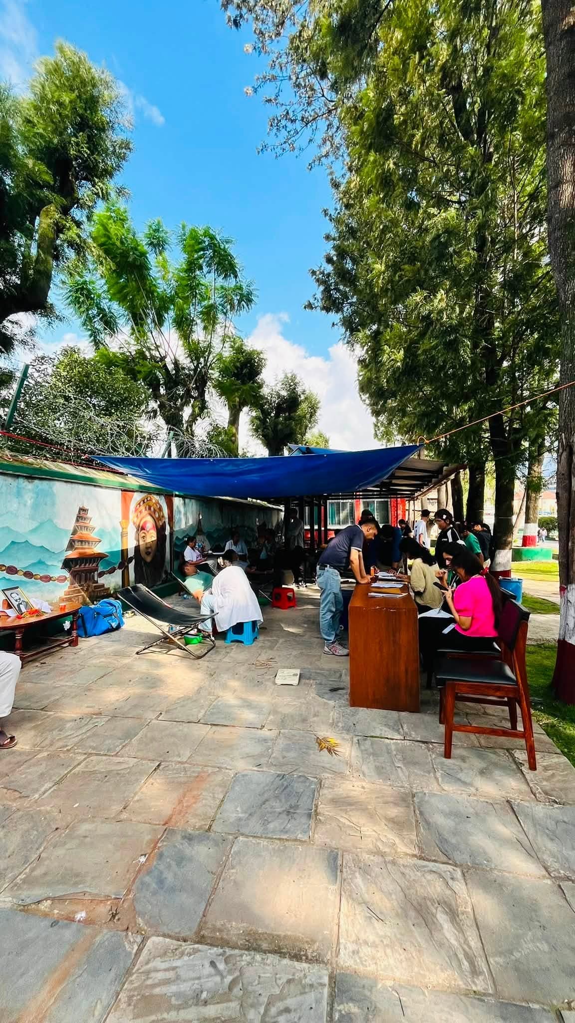 People gather under a blue tarp for an outdoor event beside a colorful wall mural.