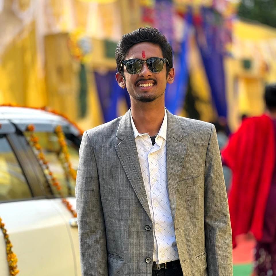 Smiling man wearing a grey plaid blazer, sunglasses, and a red tilak at an event.