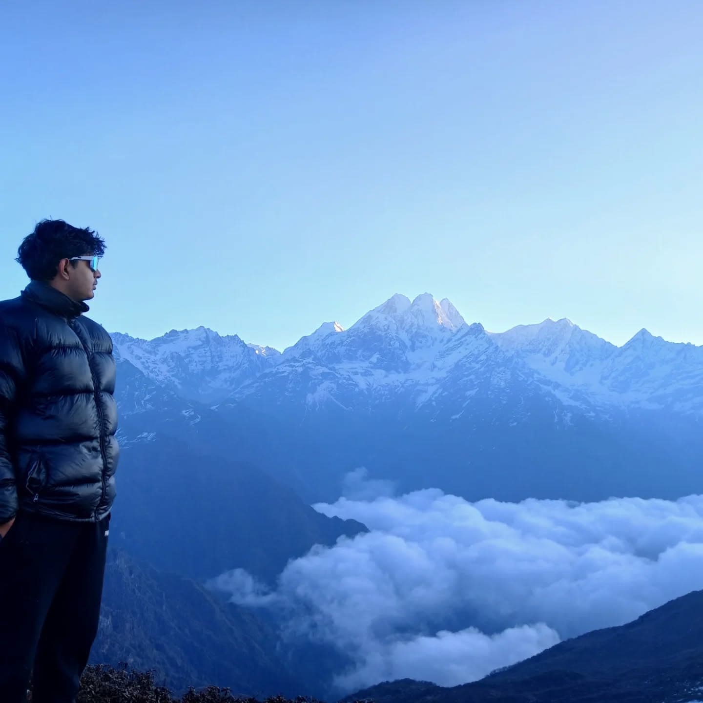 Man in black puffer jacket overlooks snow-capped mountains above a thick layer of clouds.