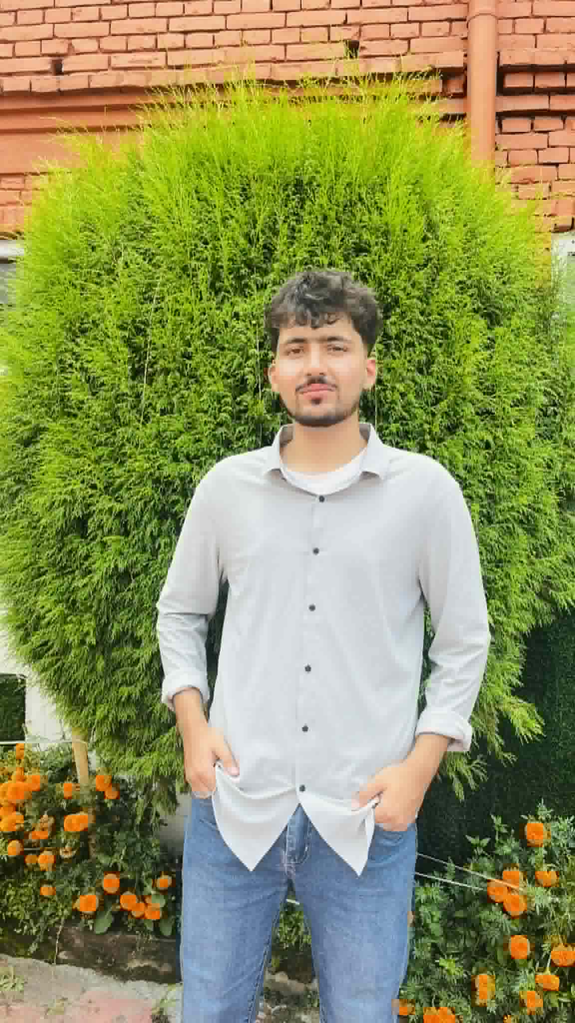 Young man in a grey shirt posing before a large green bush and orange flowers.