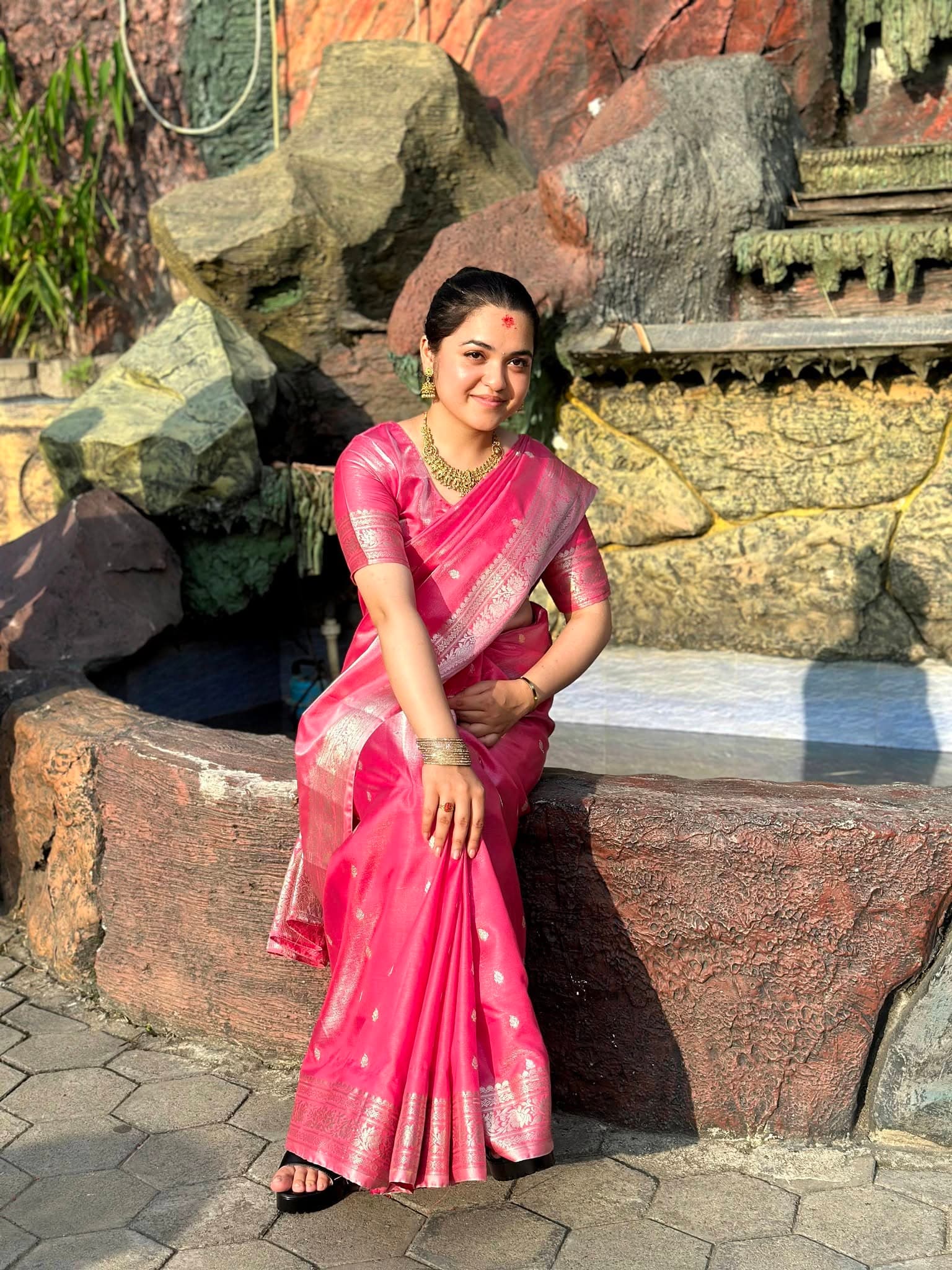 Smiling woman in a pink silk saree and gold jewelry sits on a rocky ledge.